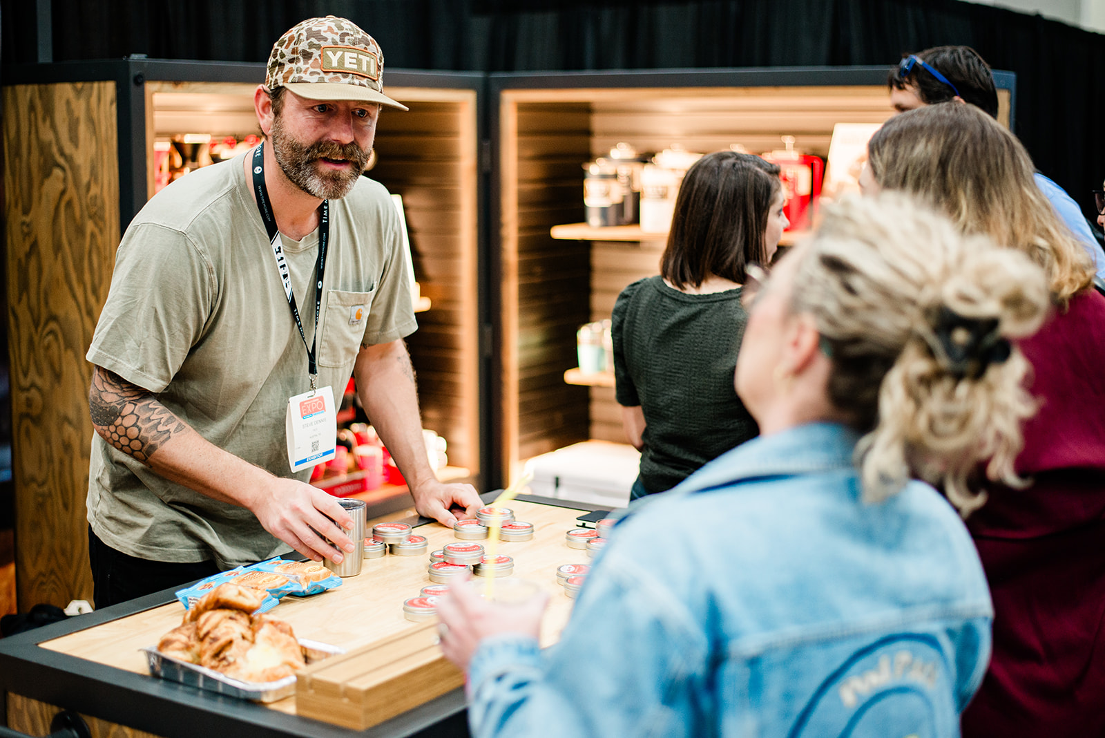 Man wearing a camo YETI hat and name badge showcasing small containers on a table to a group of people at an indoor event.