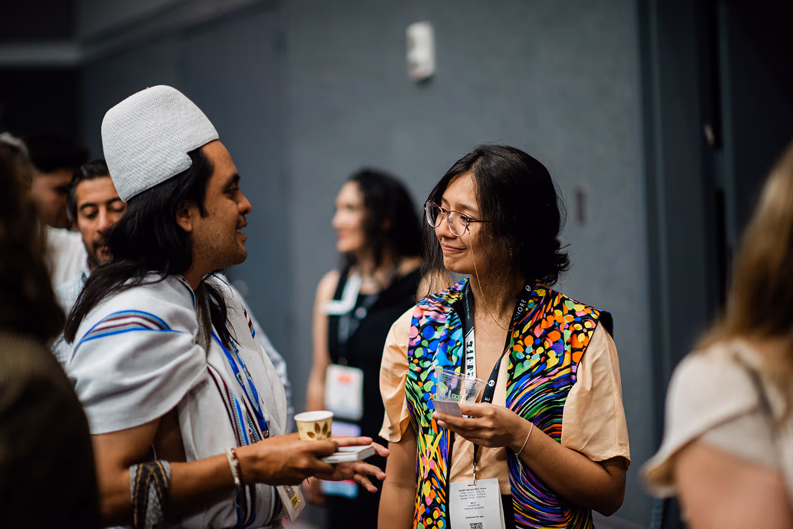 Two people engaging in conversation during an indoor event, one wearing a colorful vest and glasses, the other wearing a traditional white garment and hat.