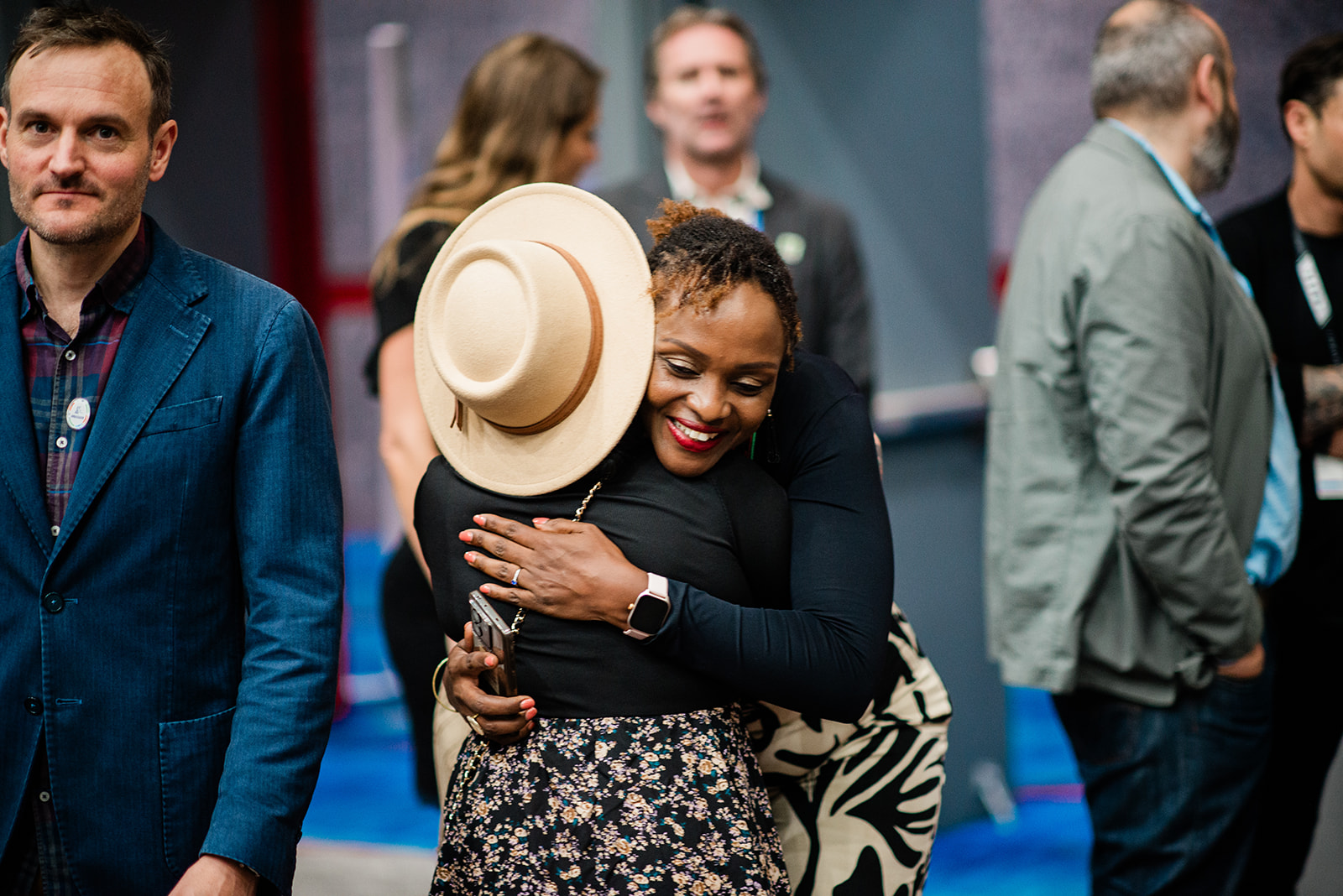 Two women warmly hugging, one wearing a beige hat and floral skirt, the other smiling with red lipstick and a smartwatch.