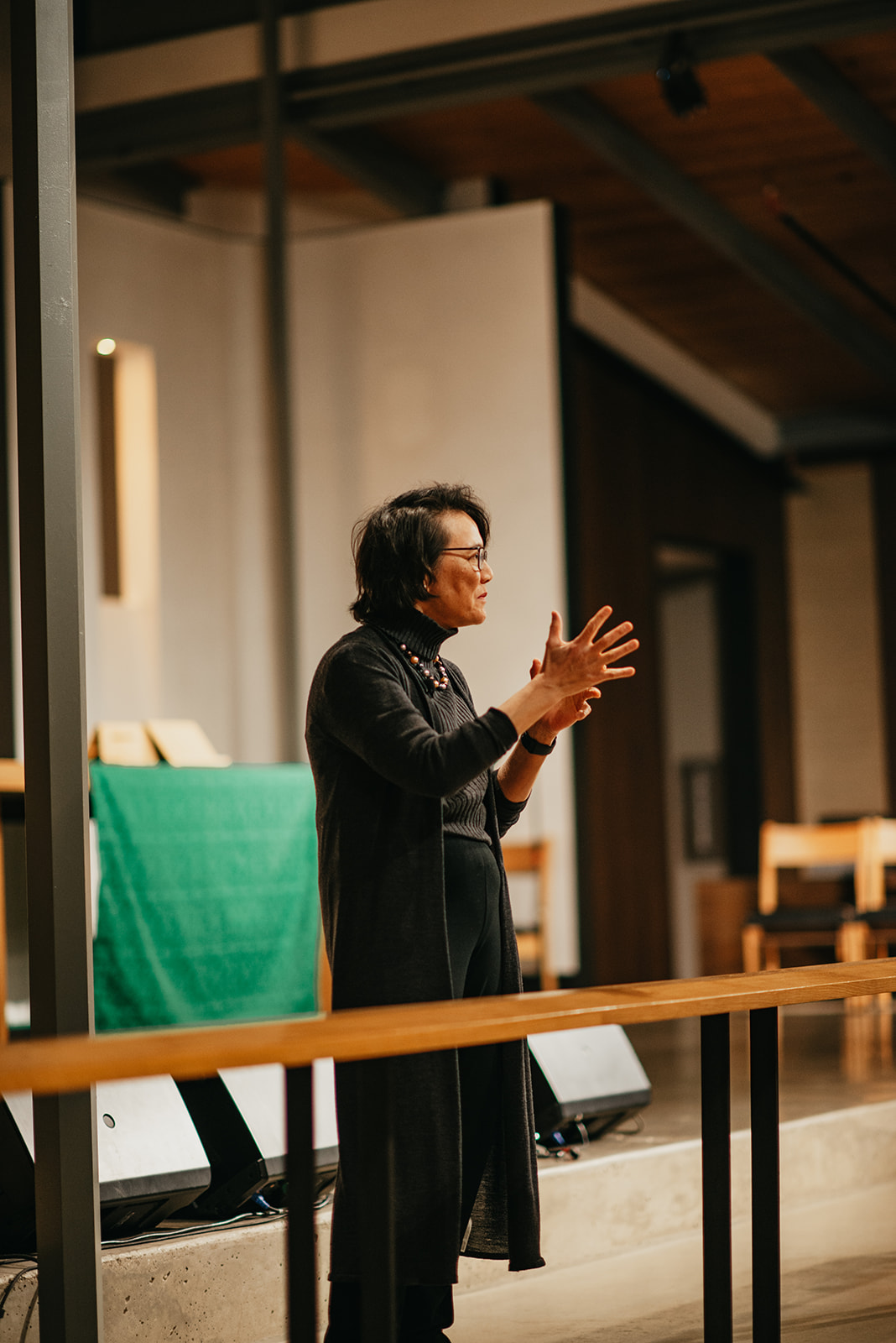 Woman wearing glasses and a dark long cardigan speaking with expressive hand gestures indoors.