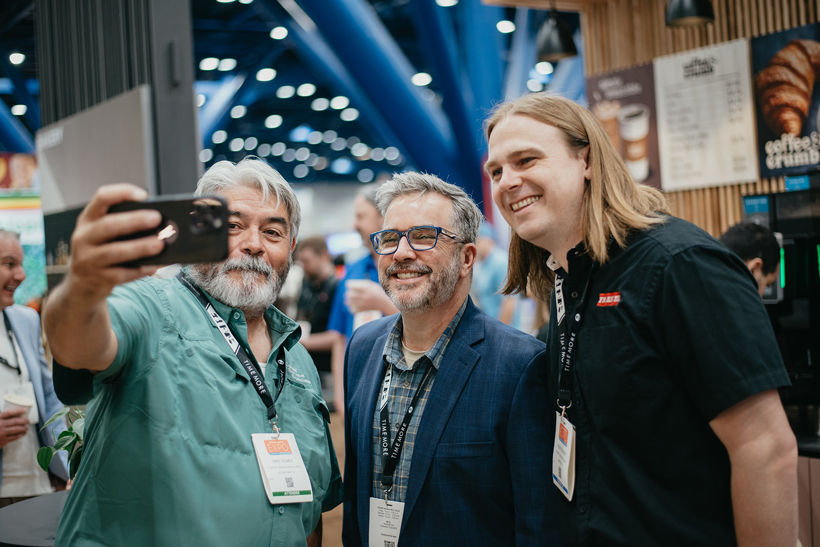 Three men smiling together as one takes a selfie at a conference or expo.