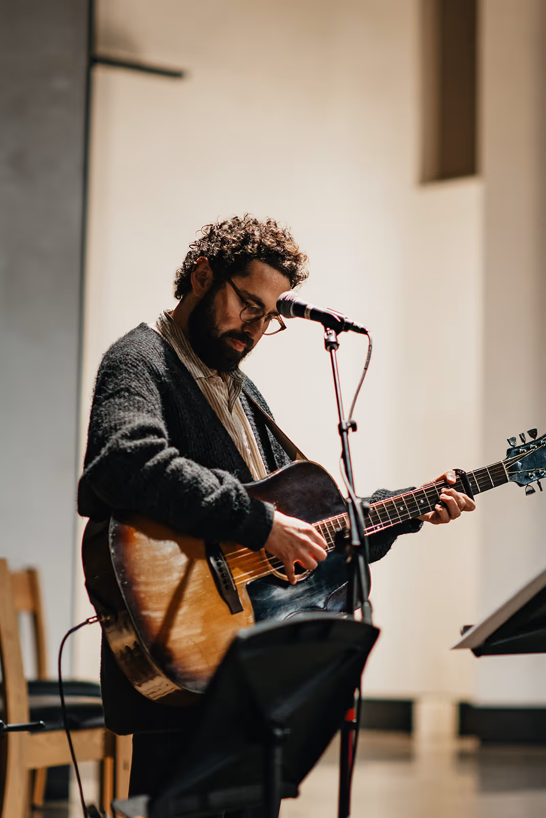Man with curly hair and glasses playing an acoustic guitar and singing into a microphone indoors.