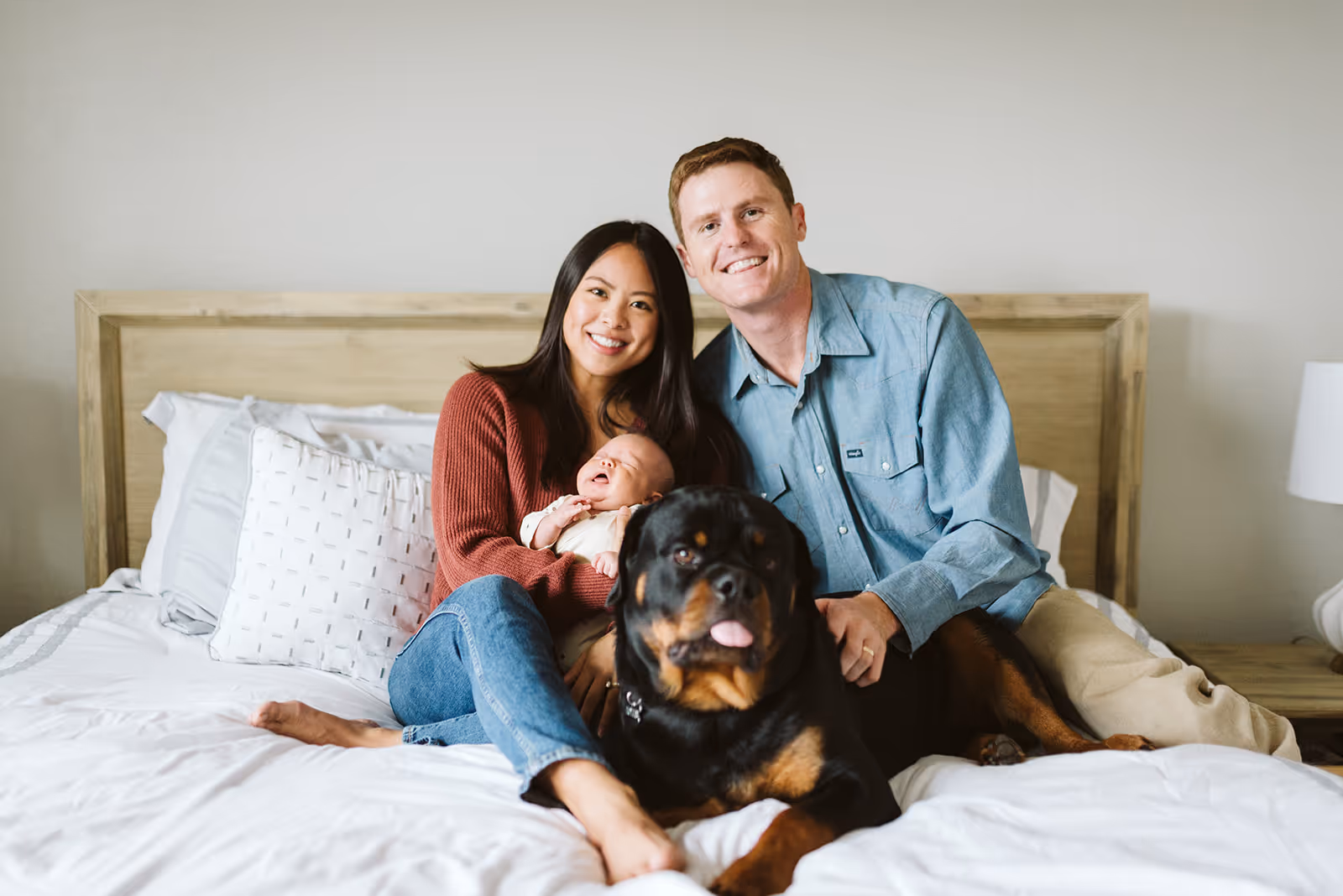 Smiling couple sitting on a bed holding a baby with a large Rottweiler dog in front of them.