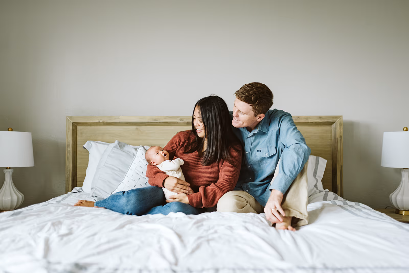 Young couple sitting on a bed smiling at their baby held in the mother's arms.