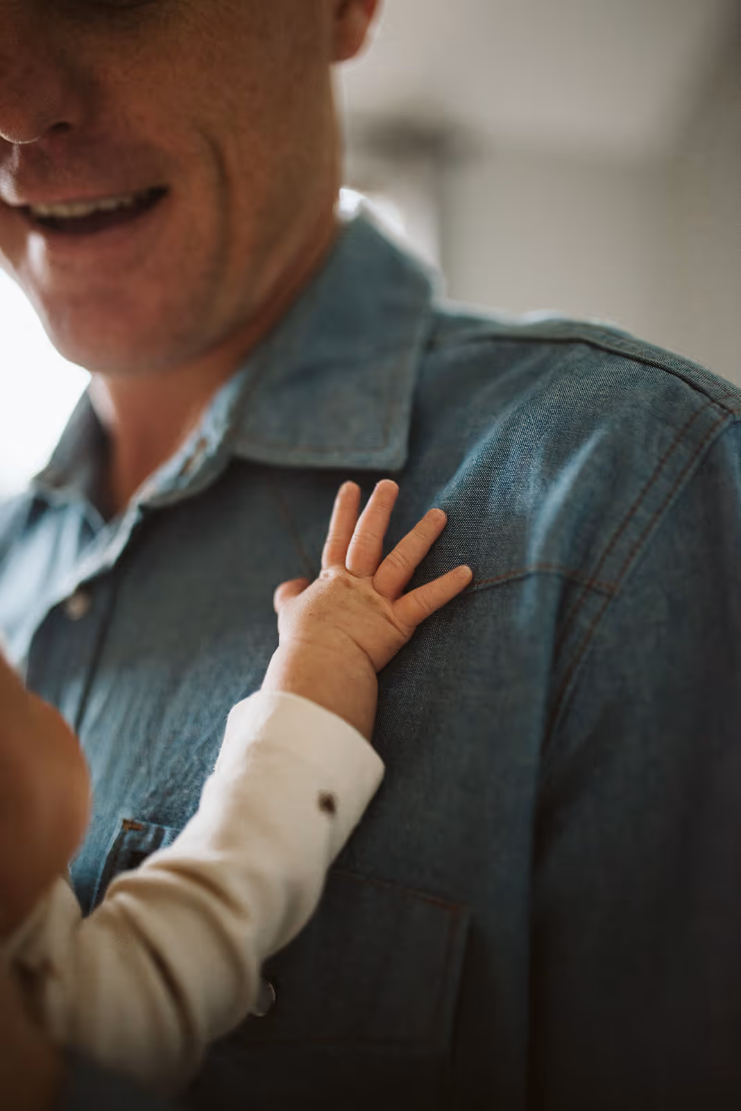 Close-up of a baby’s hand resting on a man’s denim shirt shoulder.