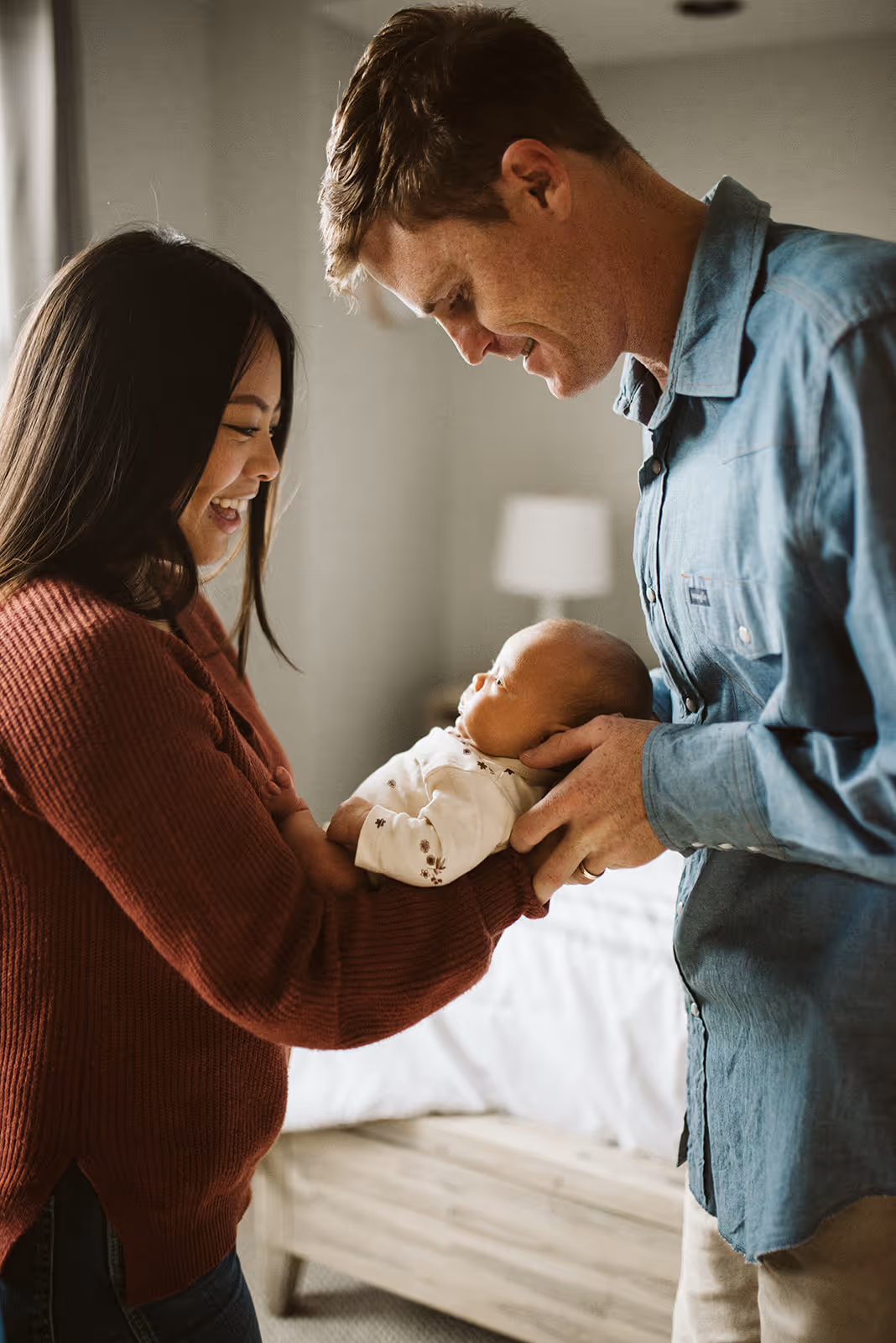 Smiling parents holding and looking lovingly at their newborn baby in a cozy bedroom.
