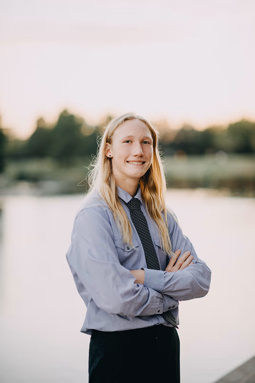 a woman with long blonde hair wearing a tie and standing with her arms crossed
