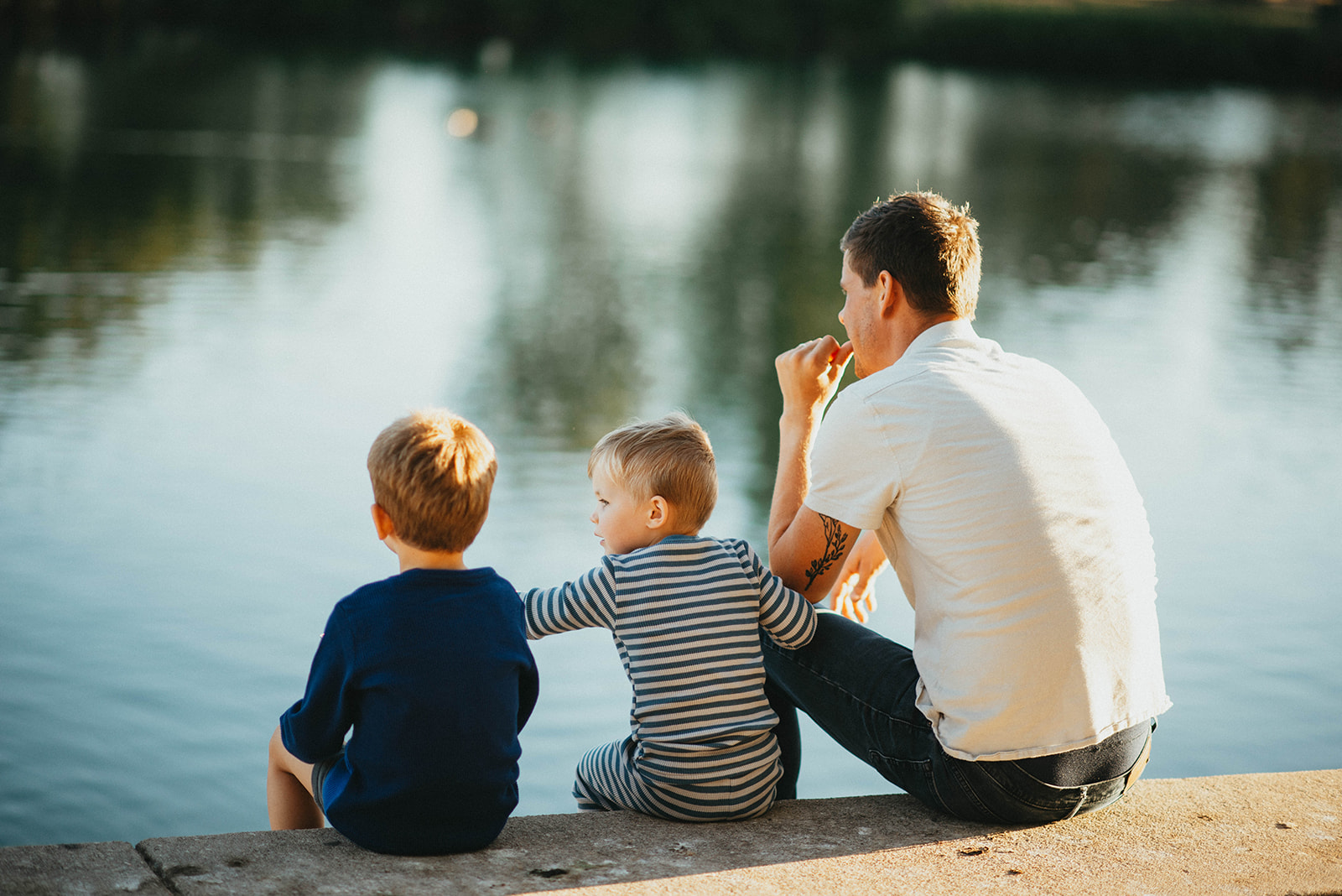 Man and two young boys sitting on a concrete ledge by a calm lake, facing the water during daylight.