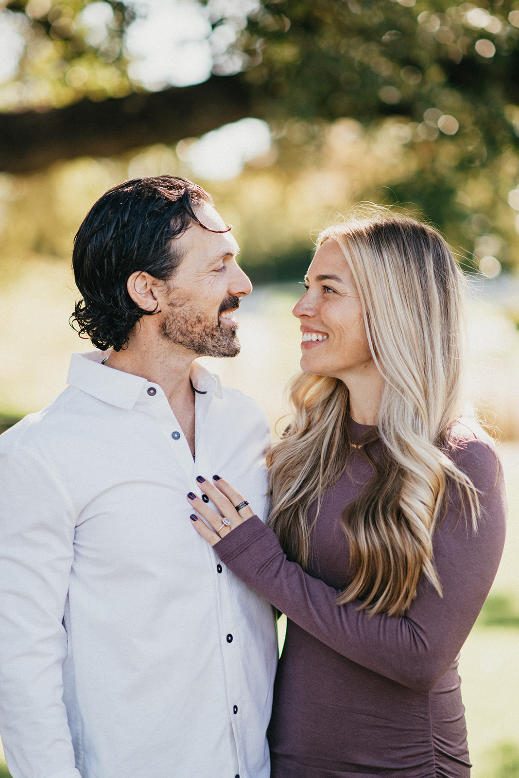 Smiling couple outdoors facing each other, the woman with long blonde hair wearing a purple top and the man with dark hair and beard wearing a white button-up shirt.