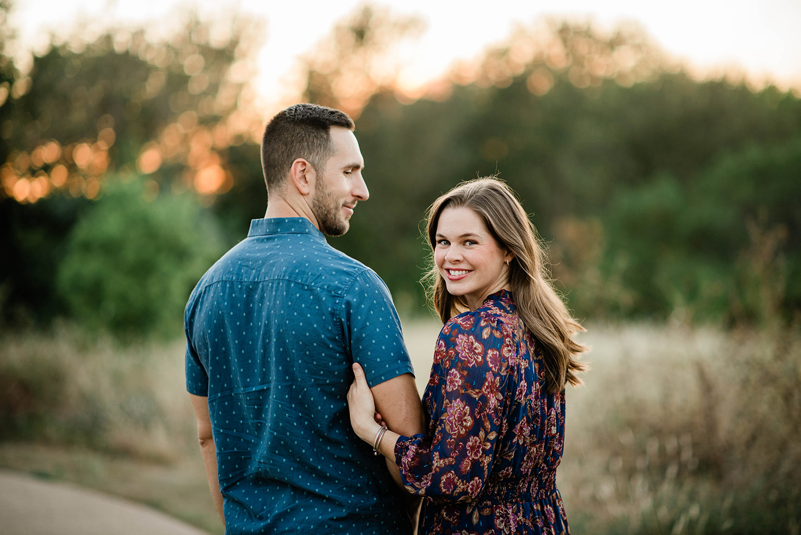 Smiling woman in floral dress holding arm of man in blue shirt outdoors at sunset.