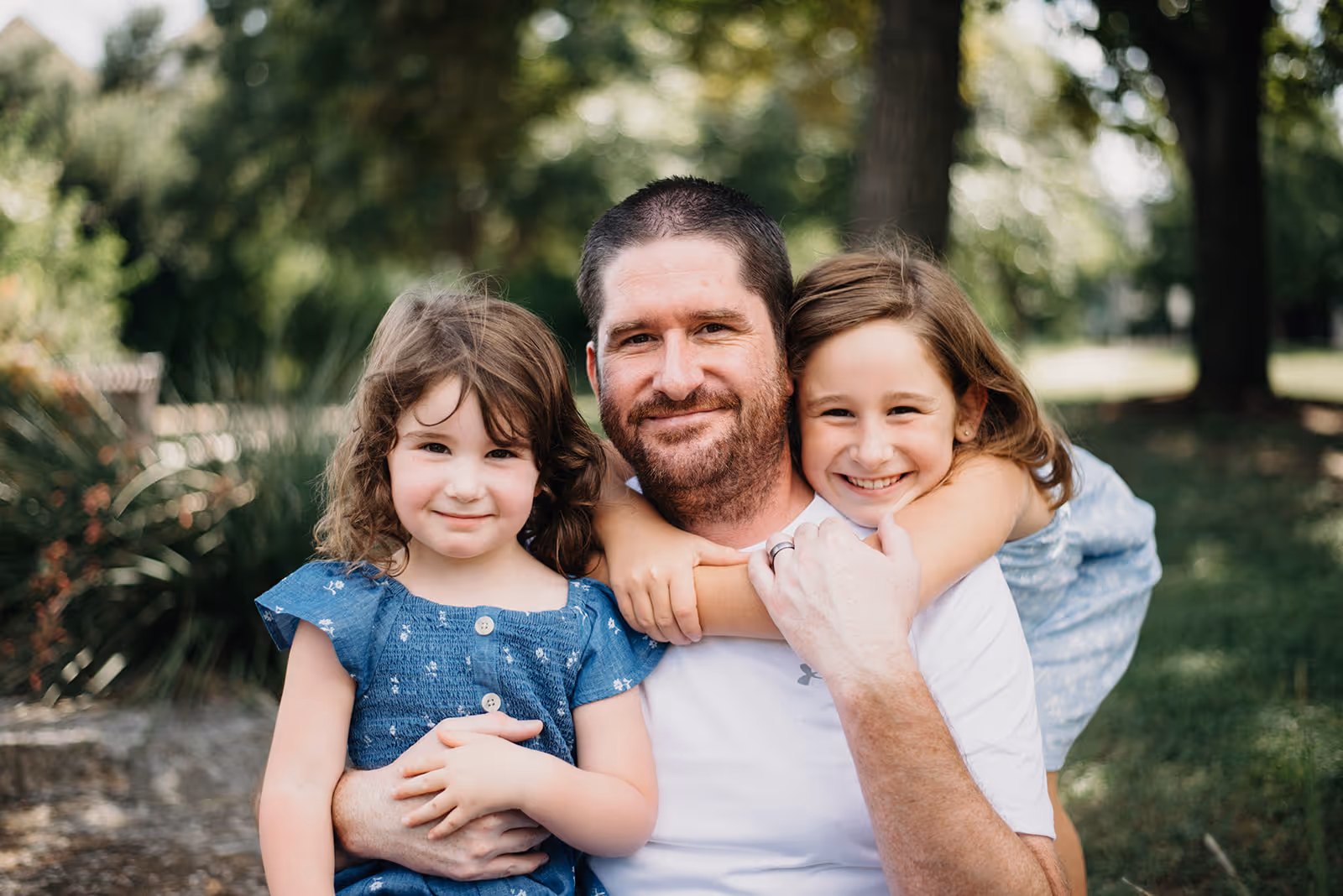 Man with beard hugging two smiling young girls outdoors with trees in the background.