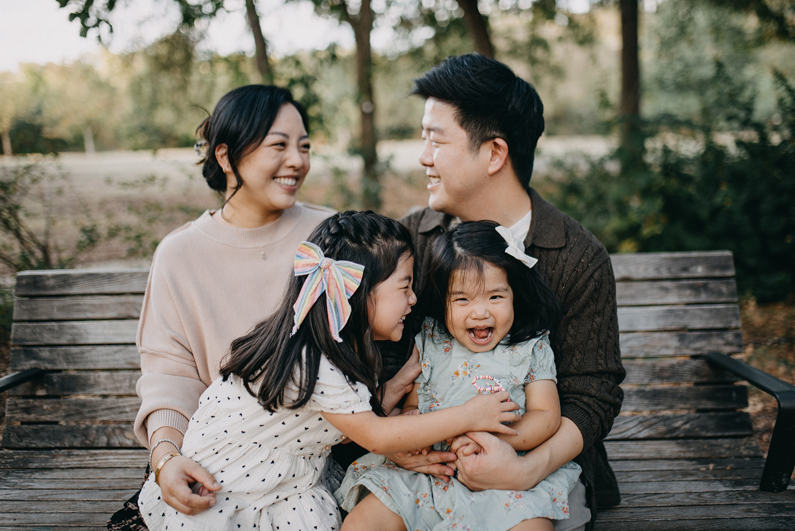 Smiling family of four with two young daughters sitting closely together on a wooden bench in a park.