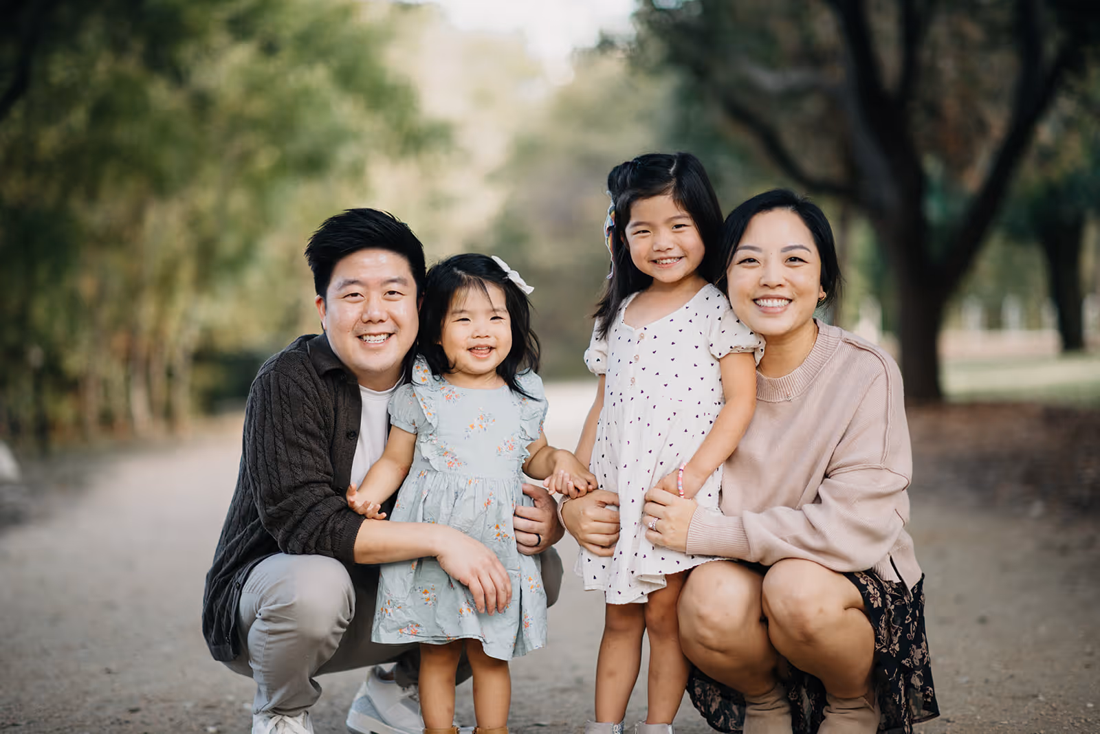 Smiling family of four with father and mother kneeling beside two young daughters on a tree-lined path.