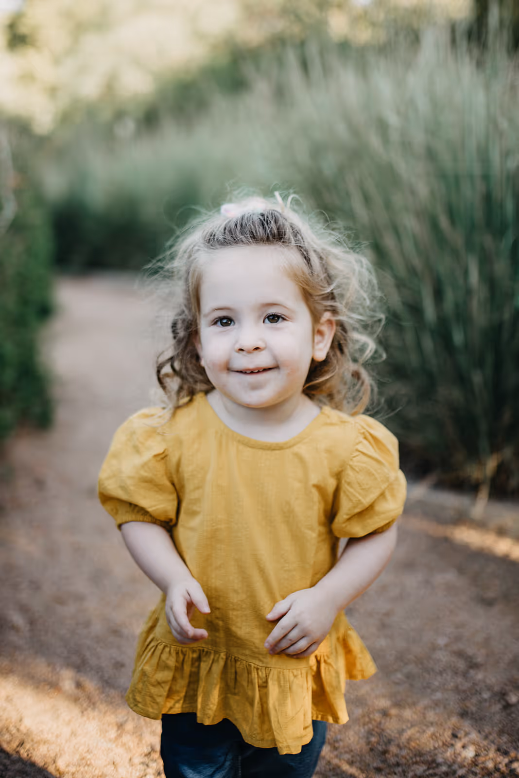 Smiling young child with curly hair wearing a yellow dress standing on a dirt path outdoors.