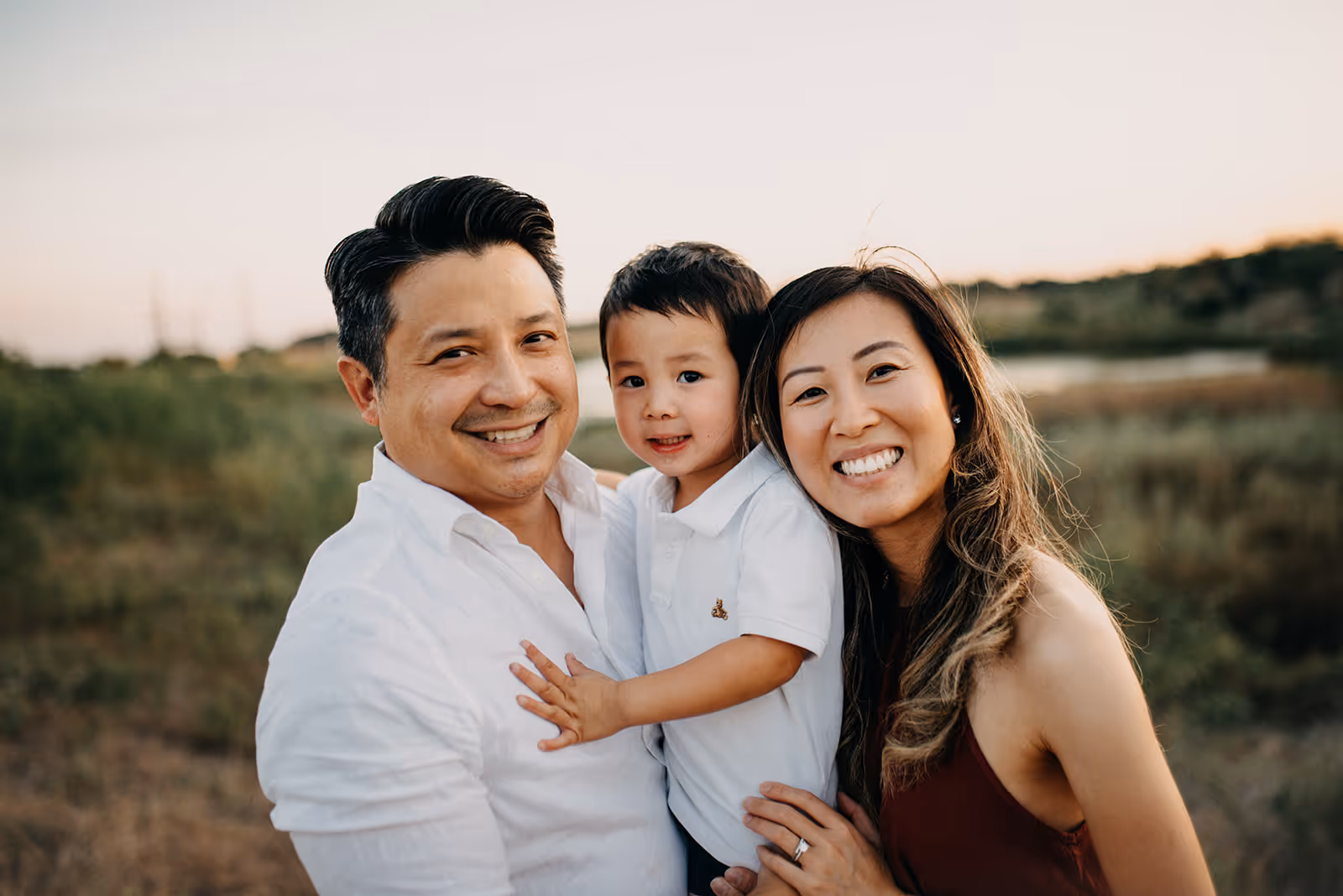 Smiling family of three outdoors during sunset with father holding a young boy and mother beside them.