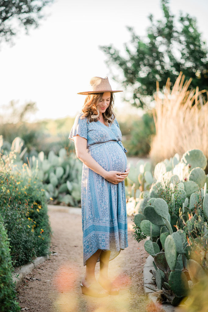 Pregnant woman in a blue patterned dress and straw hat standing on a garden path surrounded by cacti and greenery, gently holding her belly.