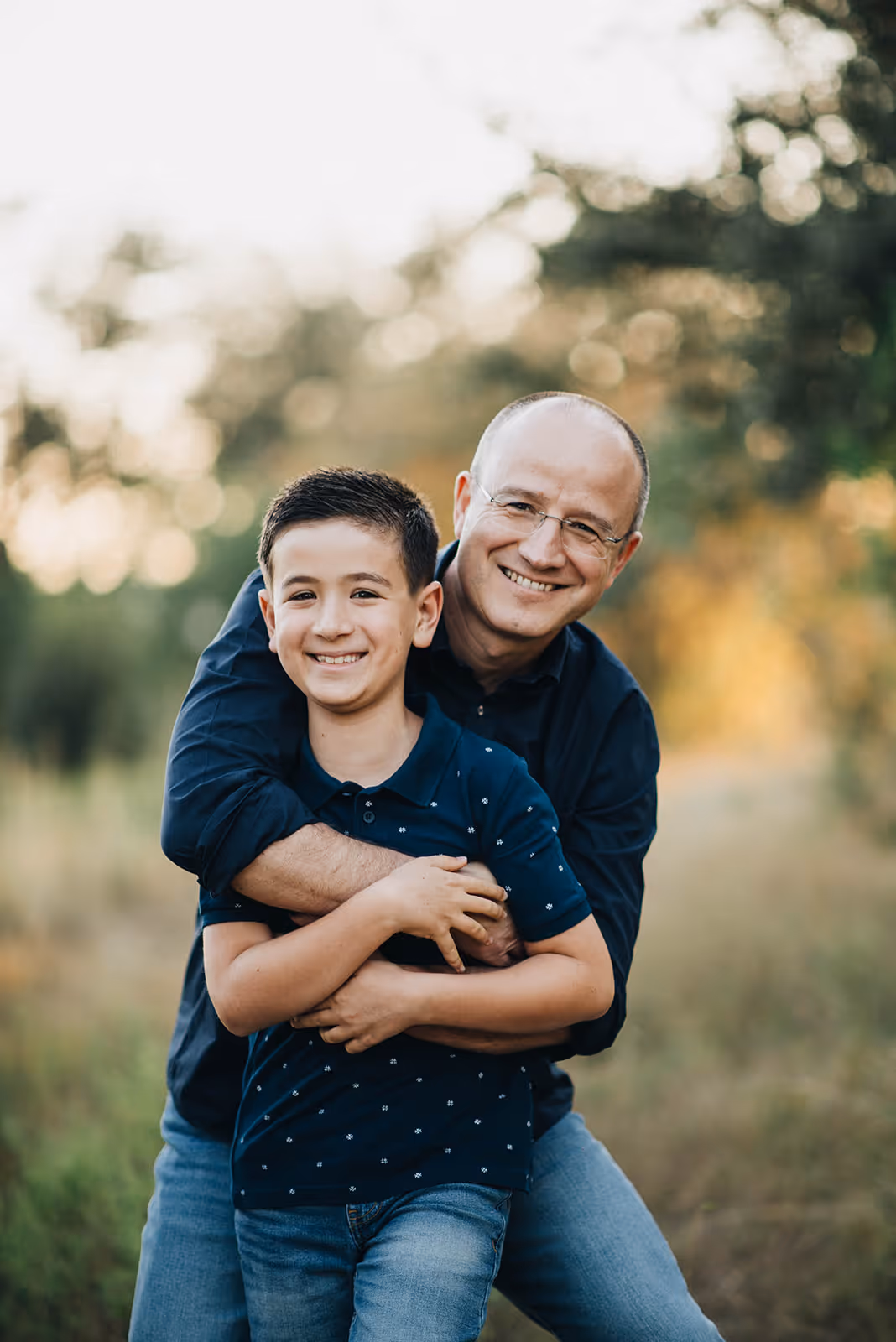 Smiling man wearing glasses hugging a smiling young boy outdoors with blurred trees in the background.