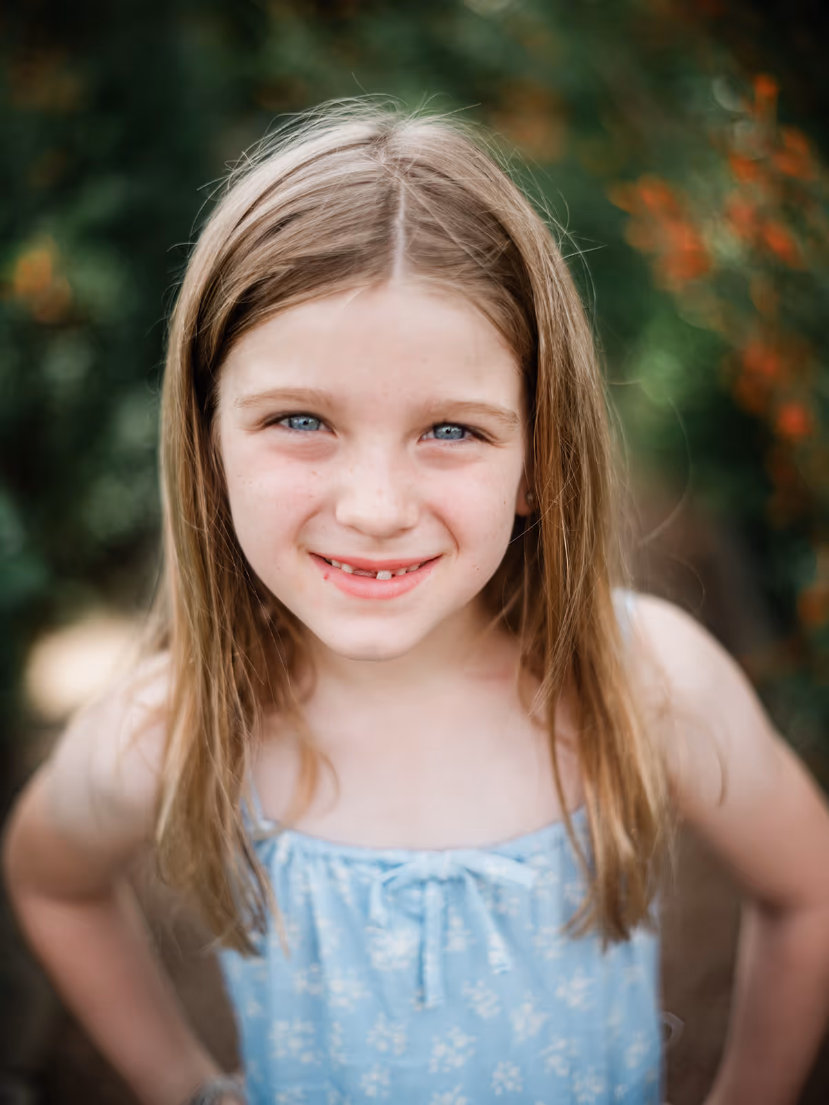 Smiling young girl with blue eyes and long light brown hair wearing a blue floral dress outdoors.