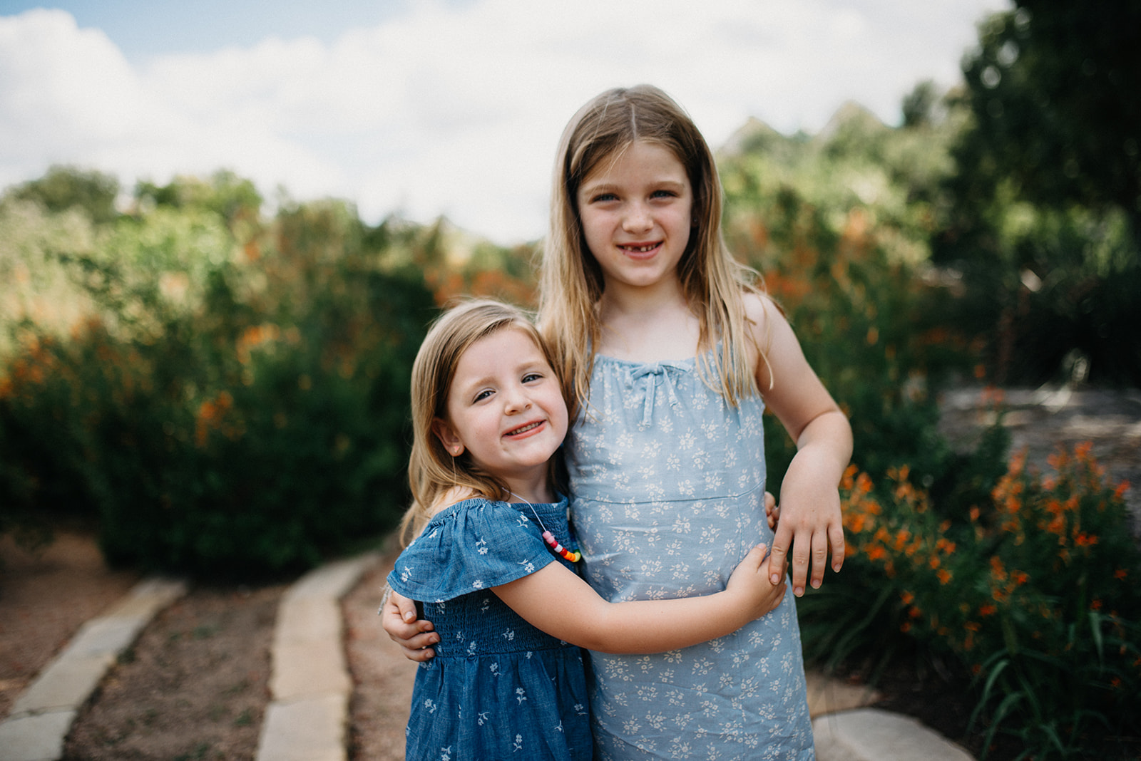 Two young girls in blue dresses smiling and hugging outdoors with green bushes and orange flowers in the background.