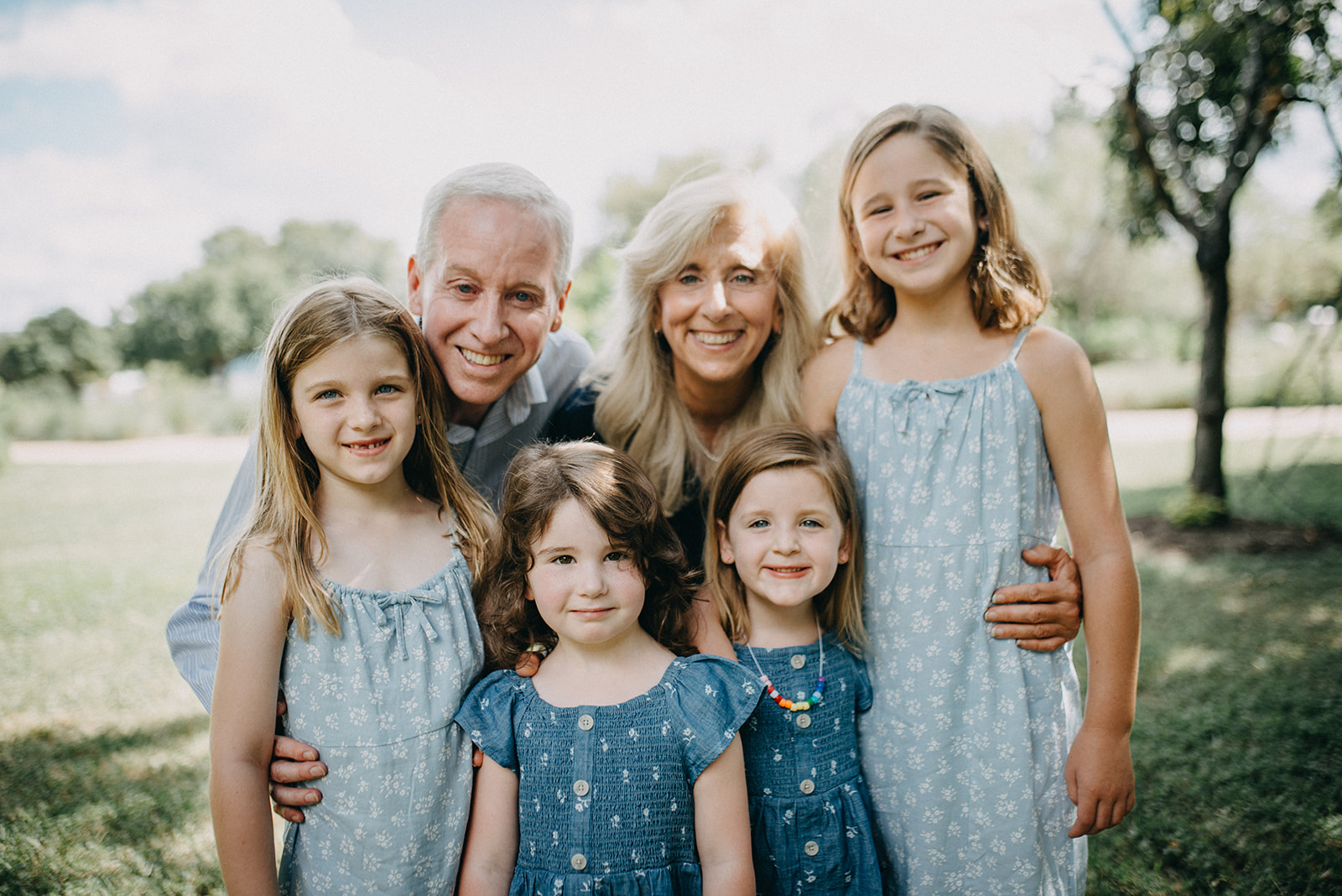 Smiling grandparents and four young girls posing outdoors on a sunny day, with green grass and trees in the background.