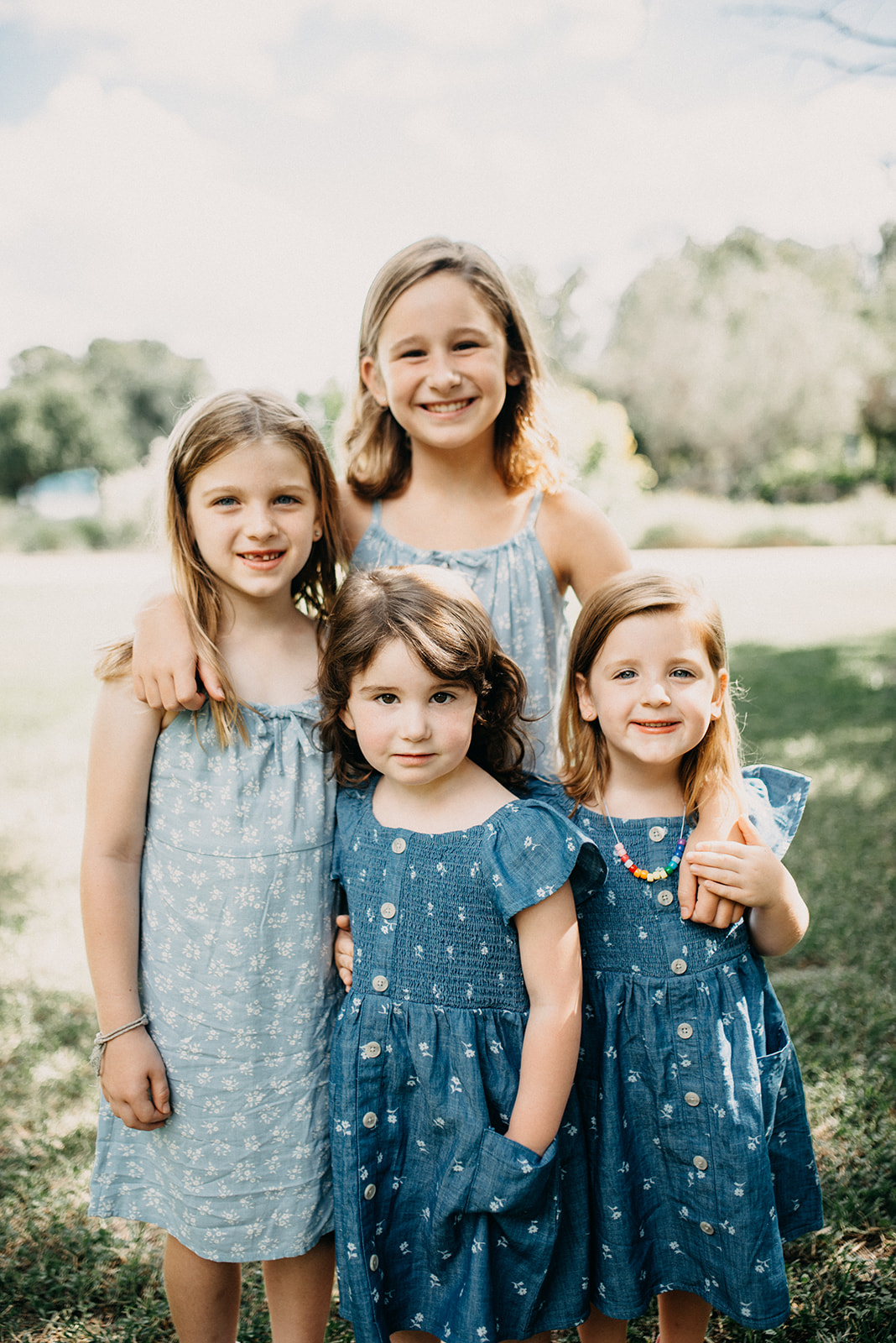 Four smiling young girls in blue dresses standing closely outdoors on grass with trees in the background.