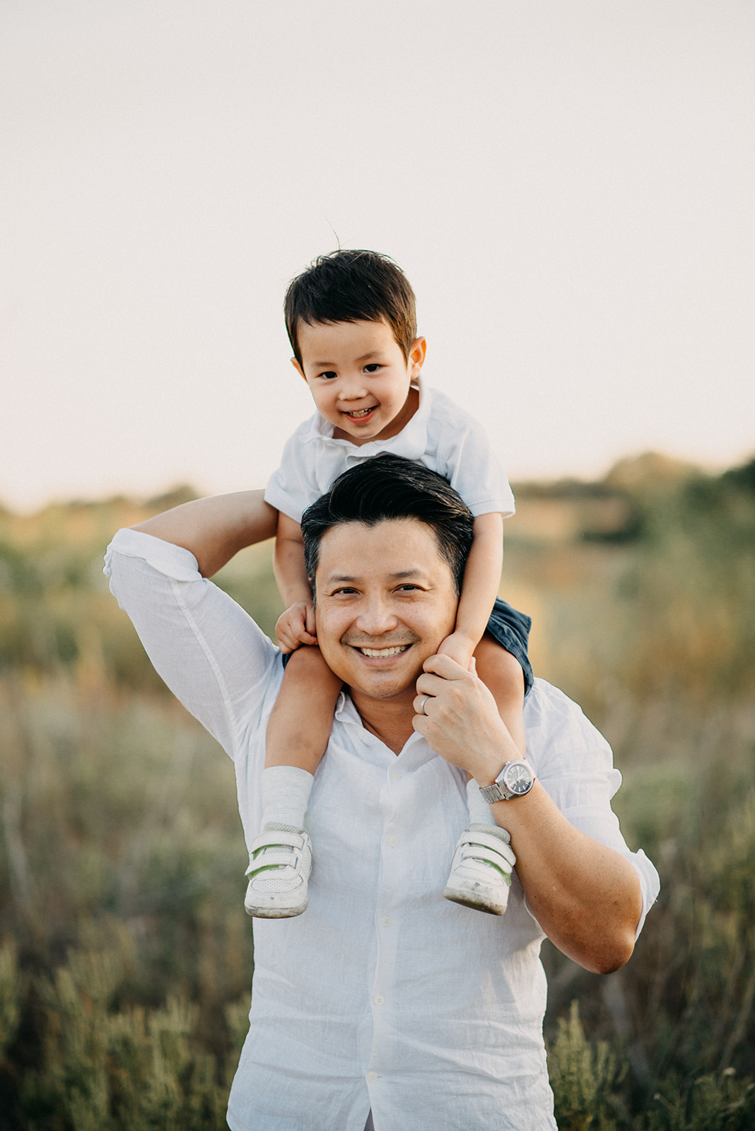 Smiling man carrying a happy young boy on his shoulders outdoors with blurred greenery in the background.
