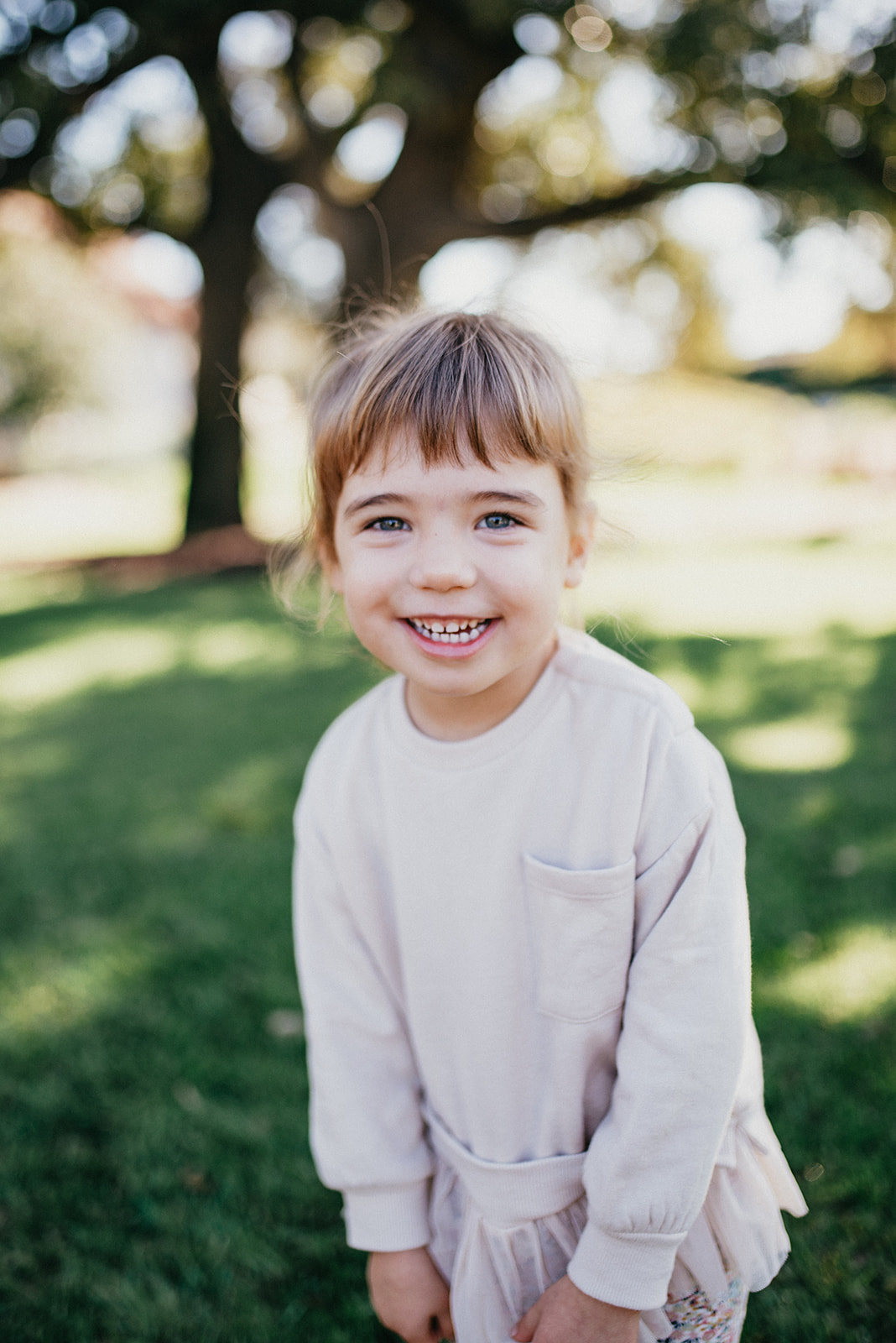 Smiling young girl with light brown hair and blue eyes wearing a light pink long-sleeve shirt standing on green grass.
