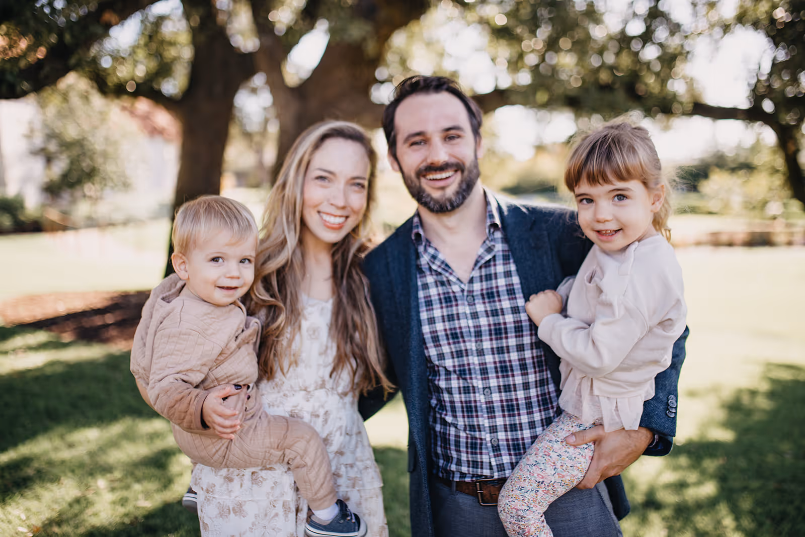 Smiling family of four outdoors with parents holding their young children under a large tree.