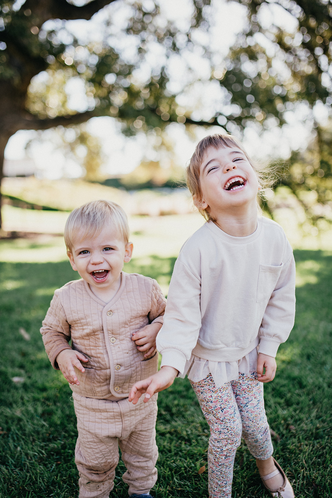 Two young children laughing and holding hands outdoors on green grass with trees in the background.