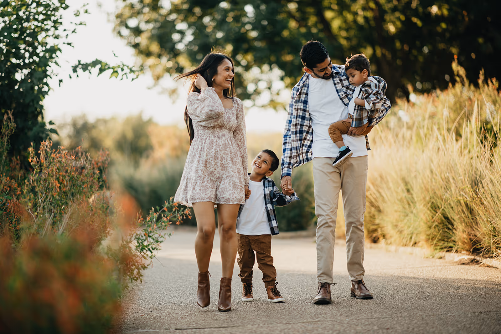 Smiling family of four walking outdoors on a sunny day, surrounded by green trees and plants.