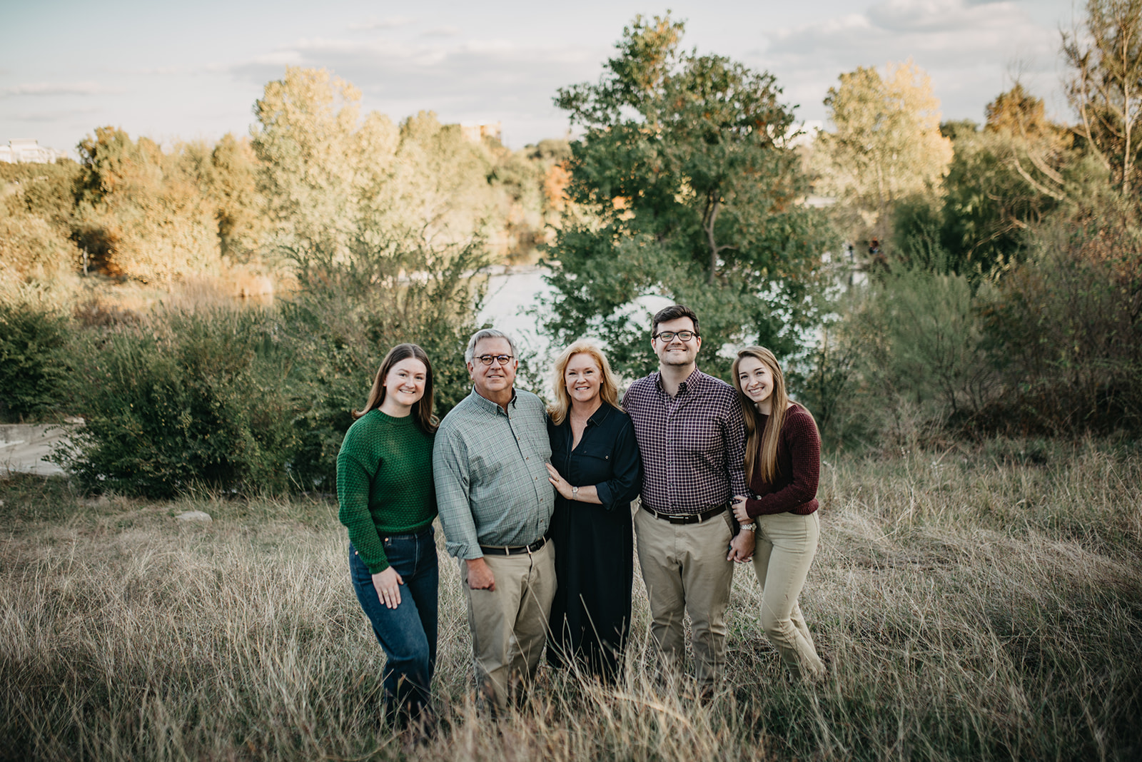 Five people standing closely together in a grassy field with trees and a body of water in the background, smiling at the camera.