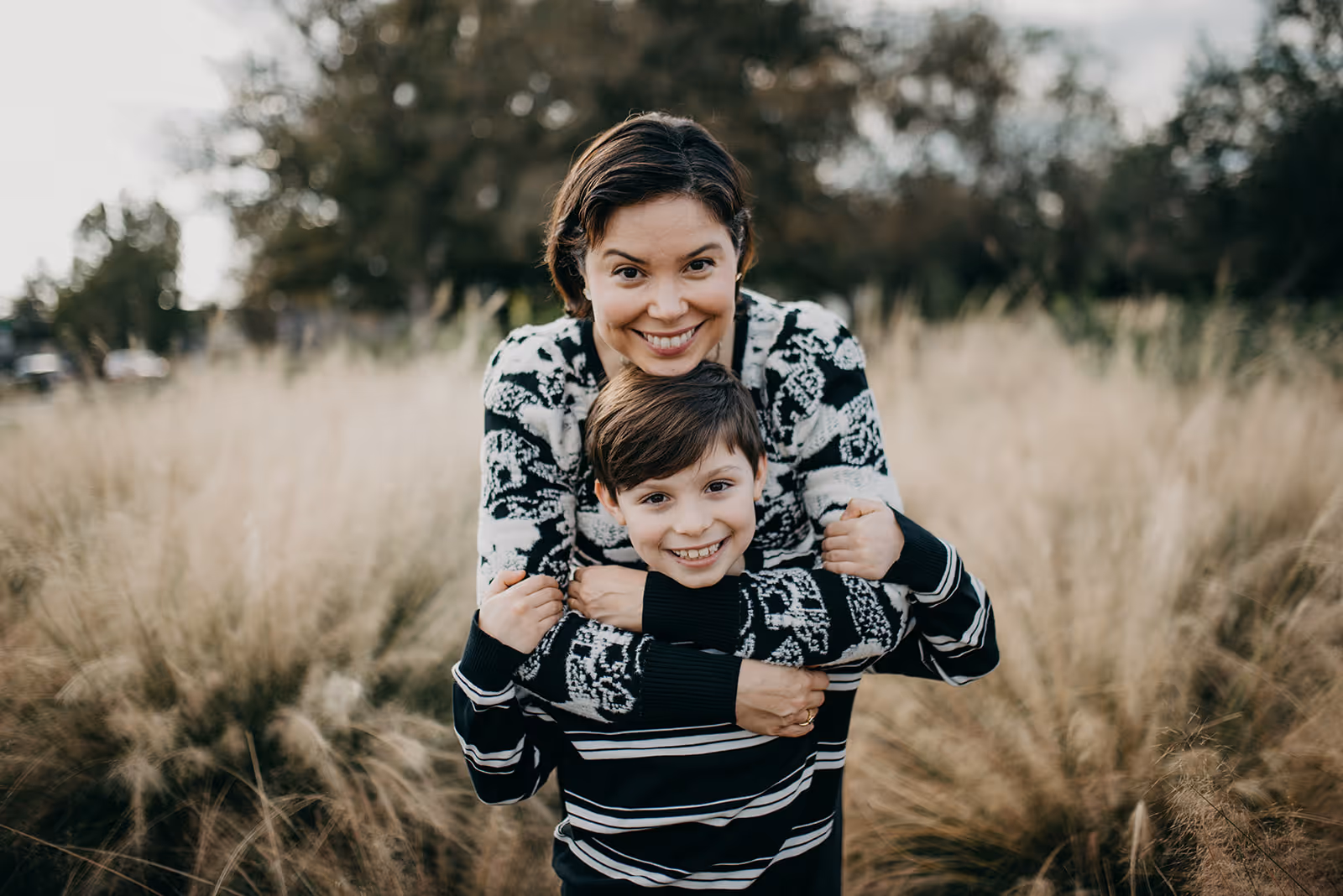 Smiling woman hugging a smiling young boy from behind in a field of tall grass.