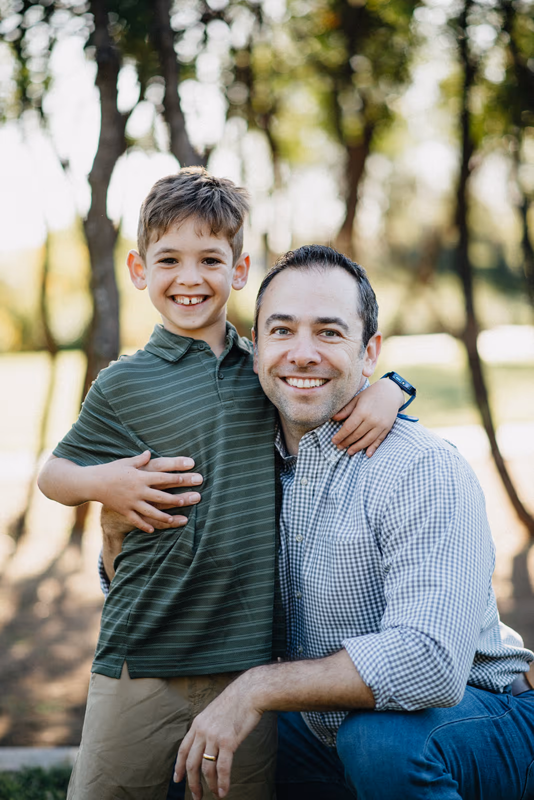 Smiling man kneeling outdoors with arm around happy young boy wearing a green striped shirt.