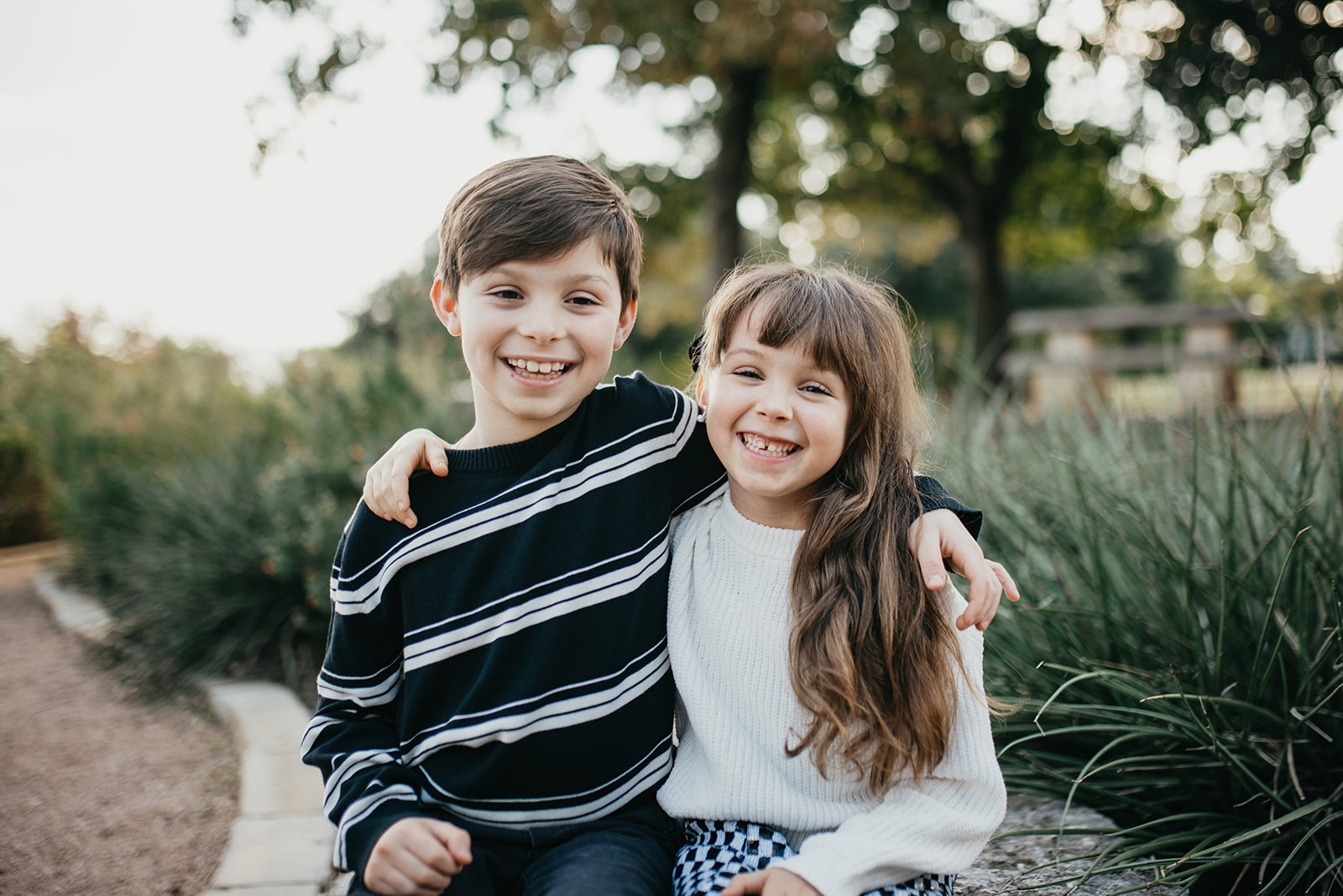 Two smiling children sitting outdoors with arms around each other, surrounded by greenery.