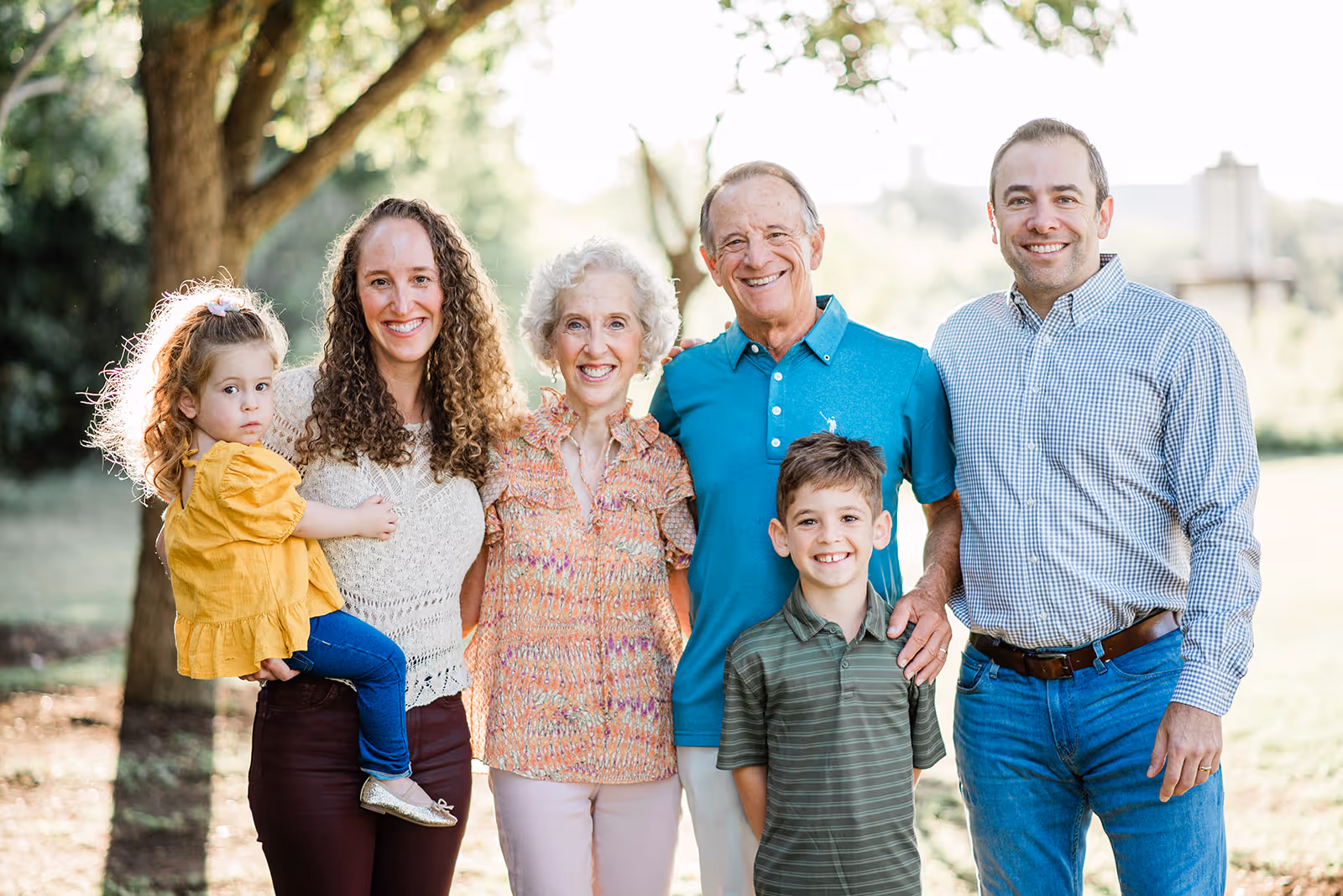 Multi-generational family in a park with a woman holding a young girl, an elderly couple, a young boy, and a man, all smiling except the girl.