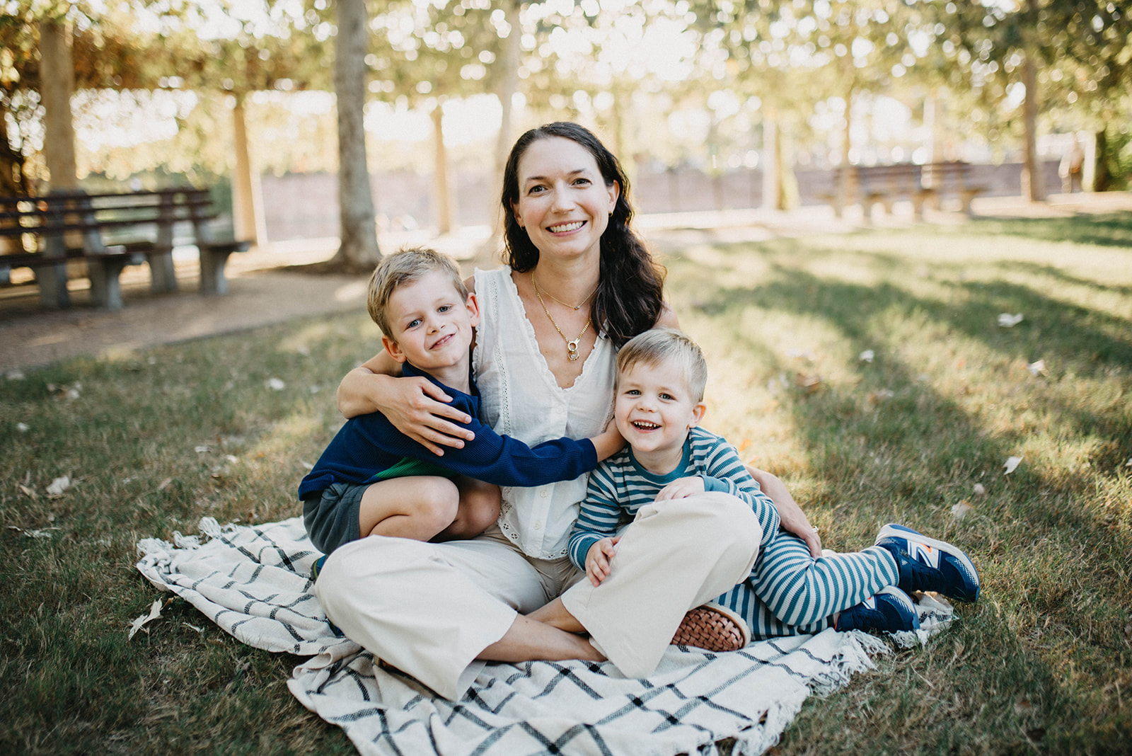 Smiling woman sitting on a blanket in a park with two young boys, one hugging her and the other leaning against her leg.