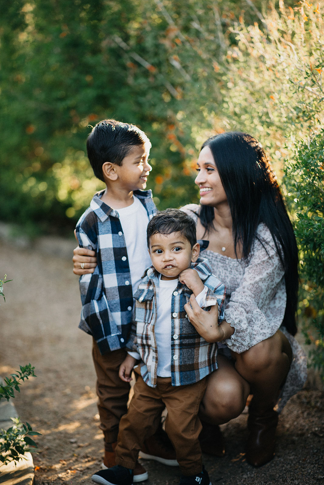 Smiling woman crouching and hugging two young boys outdoors with green foliage in the background.