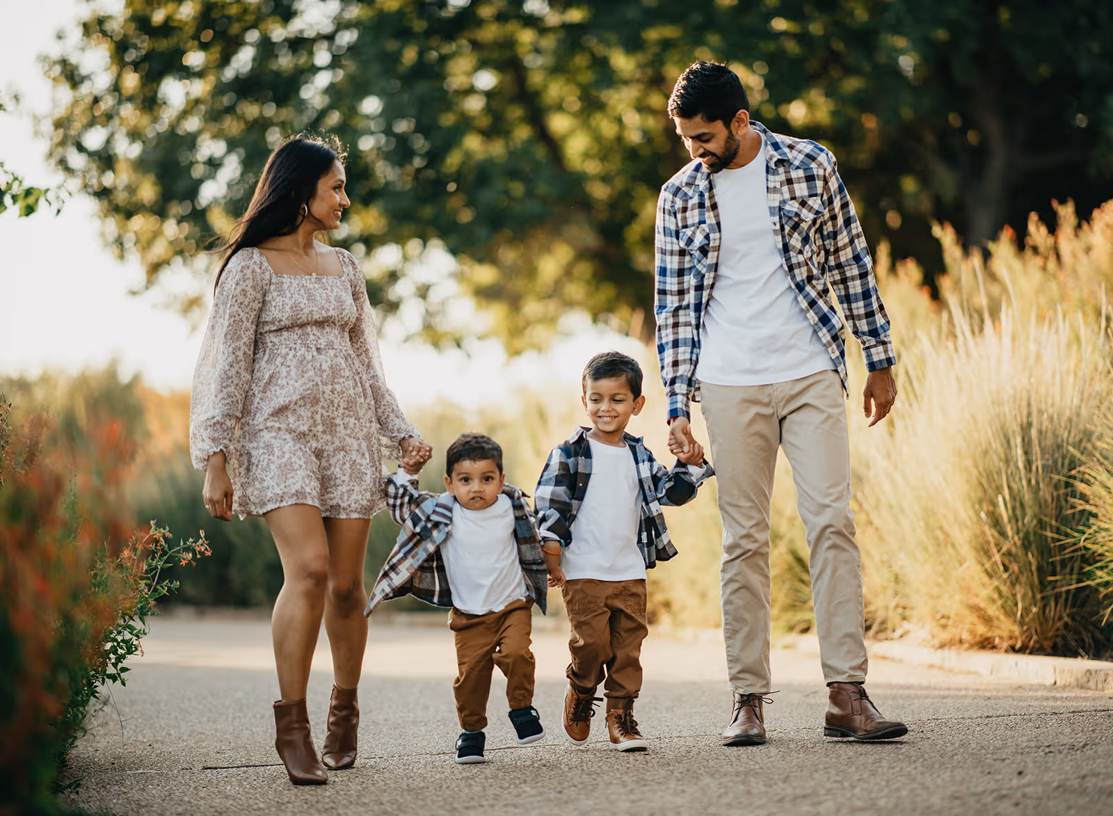 A smiling family of four walks outdoors on a path with greenery; the mother and father hold hands with two young boys between them.
