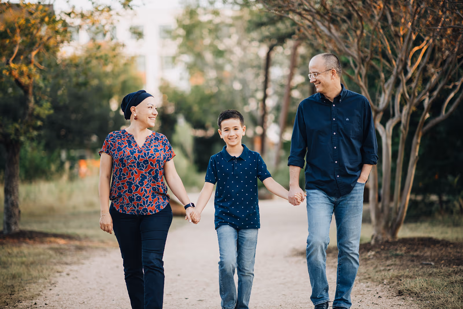 Smiling family of three holding hands and walking on a tree-lined path outdoors.
