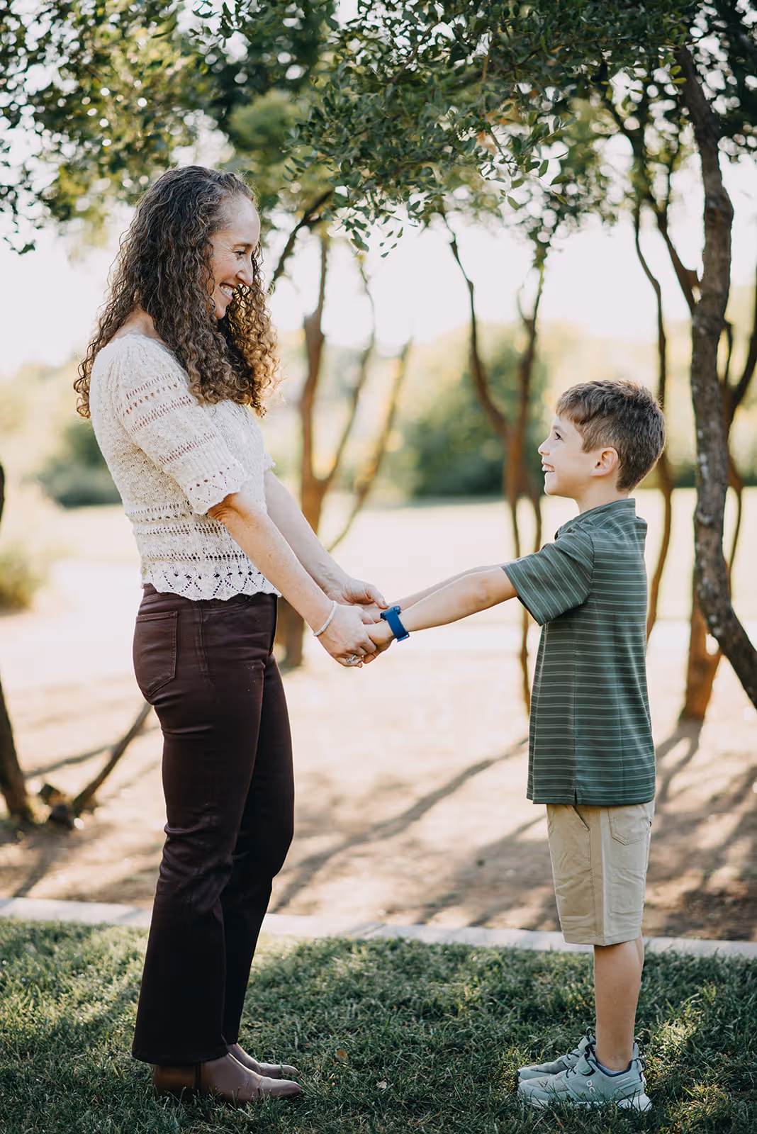 Woman and young boy holding hands and smiling at each other outdoors near trees.