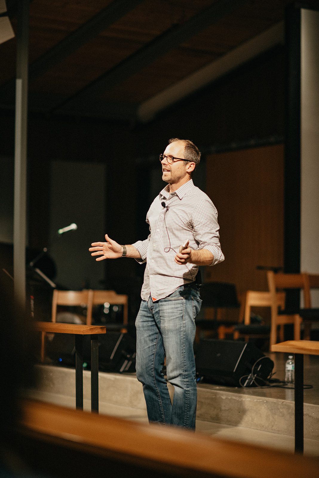 Man wearing glasses and a checkered shirt speaking in front of a small audience on a stage with wooden chairs.