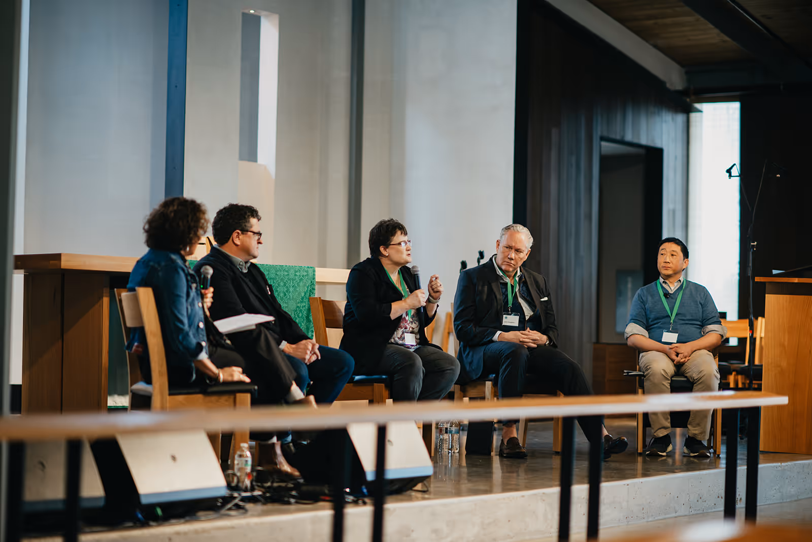 Five panelists seated on chairs in a modern indoor setting, with one woman speaking into a microphone while others listen attentively.