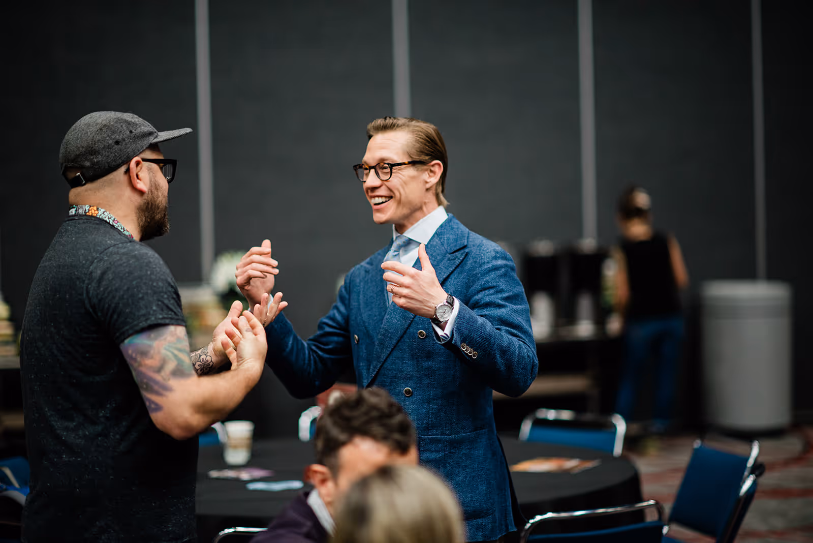 Two men conversing animatedly in a conference room, one wearing a blue suit and glasses, the other in a dark shirt and cap with visible tattoos.