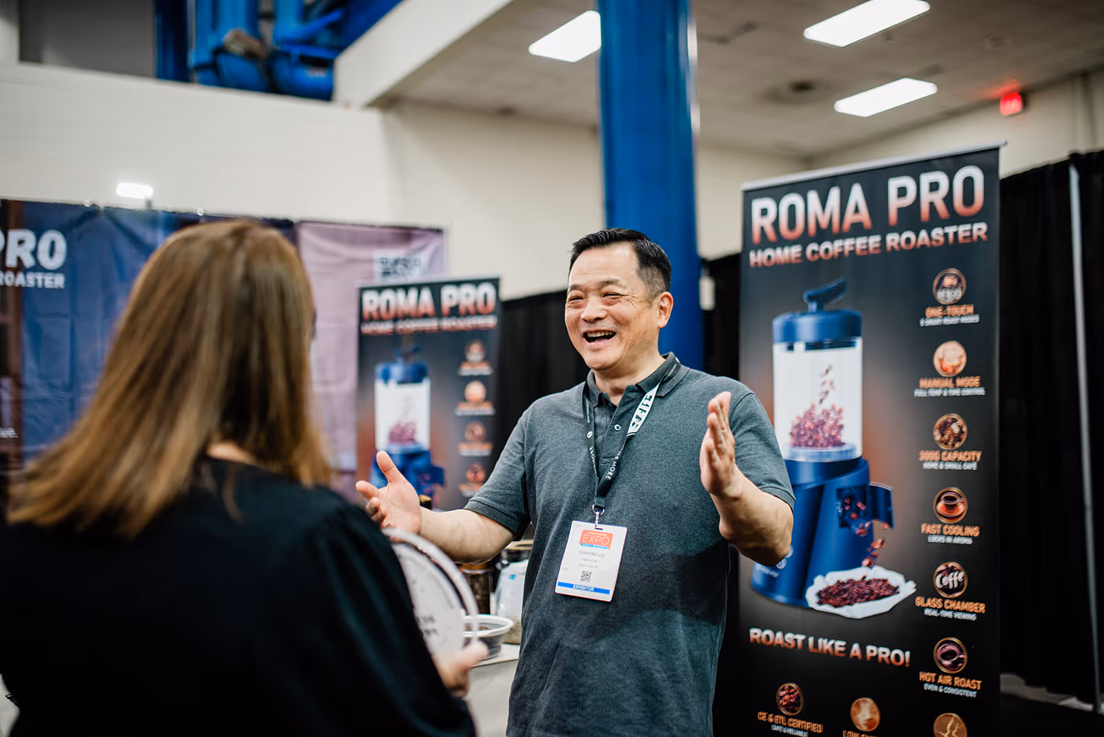 Man enthusiastically explaining the Roma Pro home coffee roaster to a woman at an expo booth.