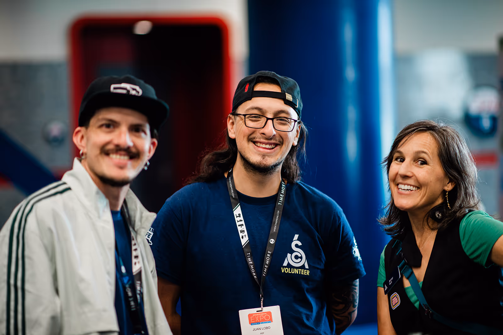 Three smiling volunteers posing indoors, one wearing a navy shirt with a volunteer logo and conference badge.