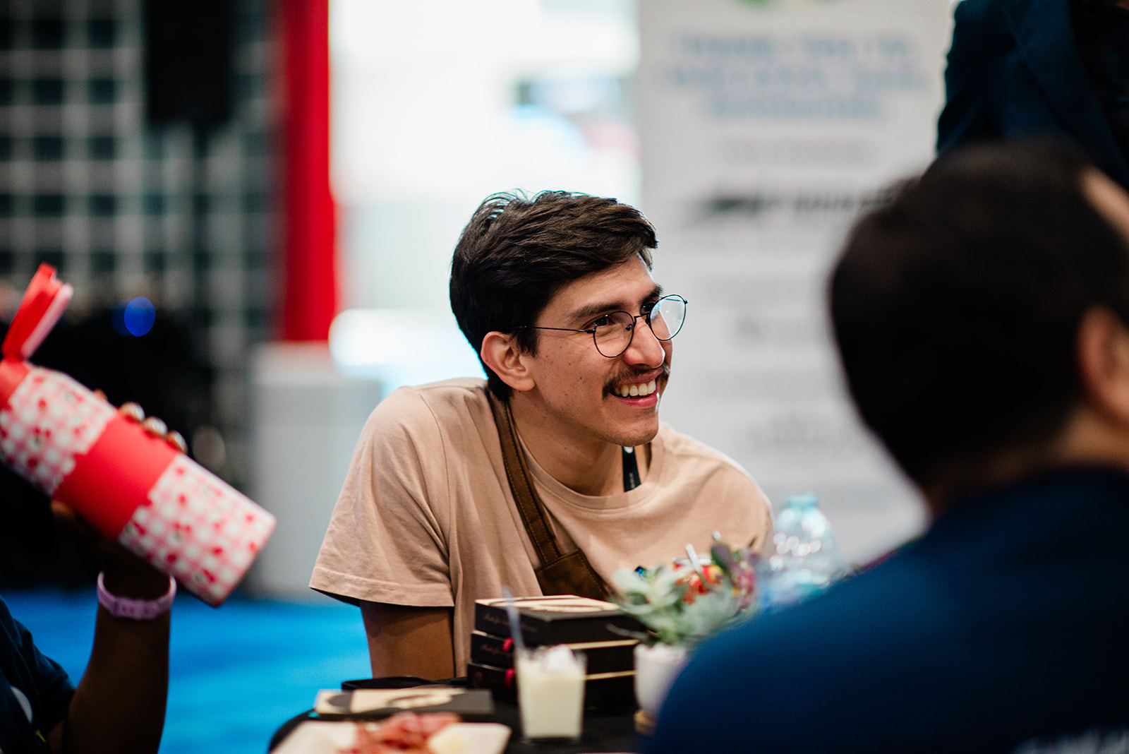 Smiling young man wearing glasses and a beige shirt sitting at a table with food and drinks.