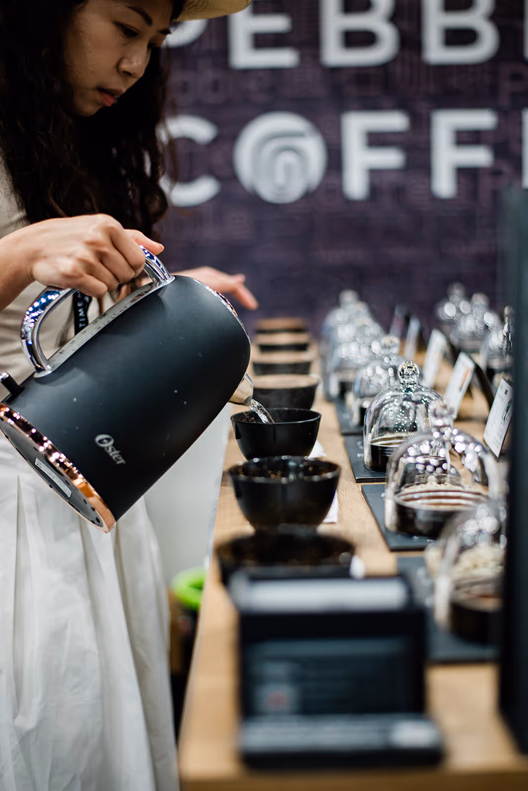 Woman pouring hot water from a black kettle into a black bowl on a wooden table with several bowls and glass containers.