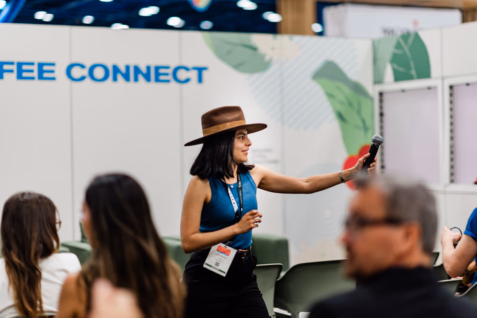 Woman wearing a brown wide-brim hat and blue sleeveless top passing a microphone to an audience member at a conference.