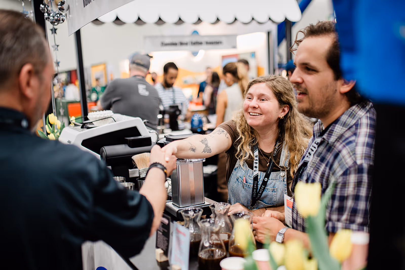 Smiling woman with tattoos and curly hair shakes hands with a man at a busy coffee booth.