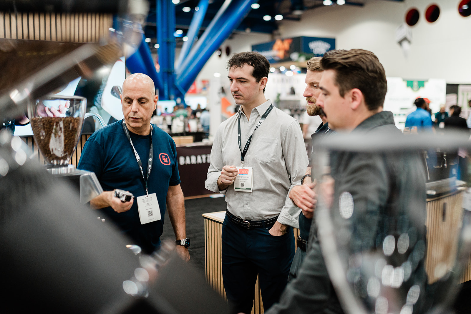 Four men wearing conference badges are engaged in discussion at an indoor expo.
